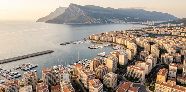 Vue aérienne réaliste de Toulon montrant le port, les immeubles résidentiels et le mont Faron sous une lumière douce.
